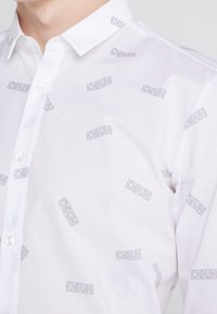 Weißes Hemd aus Baumwolle mit einem wiederholten grauen Hugo-Logo-Muster. Es verfügt über einen spitzen Kragen und eine Standard-Knopfverschluss.