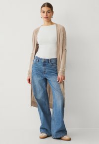 Lunga cardigan beige sopra una maglietta bianca aderente, abbinata a jeans a gamba larga blu chiaro e scarpe beige. Texture morbide e design casual.