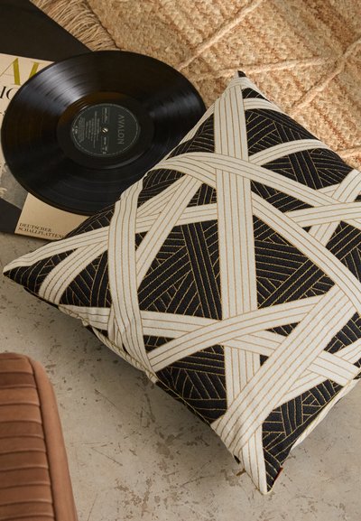 Square black pillow with white and gold geometric pattern, featuring thick woven straps and silky texture, placed near a vinyl record.