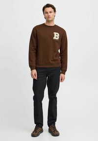 Brun sweatshirt med en teksturert bokstav "B" lapp på venstre bryst, kombinert med svarte jeans og brune treningssko.