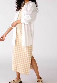Camisa blanca de botones sobre un vestido a cuadros beige con una abertura lateral, combinado con sandalias de peluche. Tejido suave, corte relajado y patrones sutiles.