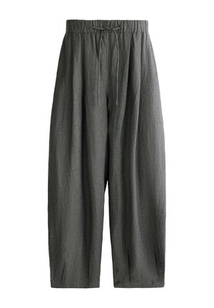 Kobieta stoi przodem, ubrana w dopasowany szary t-shirt, luźne ciemnoszare spodnie ze sznurkiem i czarne sandały na jednolitym tle.