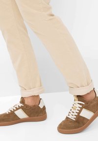Braune Wildleder-Sneaker mit weißen Akzenten, ausgestattet mit beigen Schnürsenkeln und einer strukturierten Gummisohle. Kombiniert mit hochgekrempelten hellbeigen Hosen.