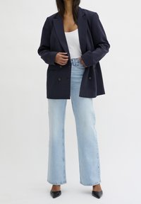 Marinblå dubbelknäppt kavaj, ljusblå high-waisted vida jeans, vit linne och svarta spetsiga högklackade skor. Slät tygstruktur.