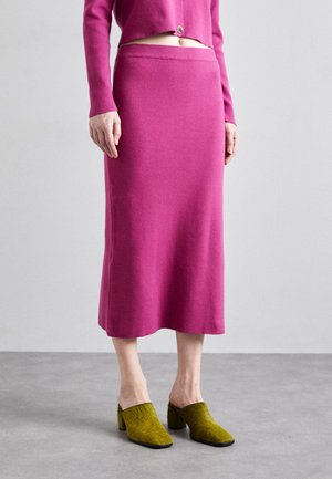 Femme portant une jupe midi rose vif avec un cardigan court assorti et des chaussures mules à talons verts, debout sur un sol gris.