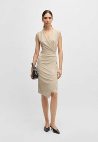 Beige wrap dress with short sleeves, gathered texture at the waist, and an asymmetrical hem. Paired with a small black handbag and black heels.