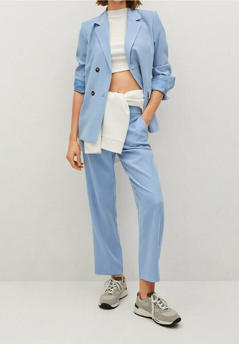Femme portant un costume bleu clair avec les manches retroussées, un top court blanc, un pull blanc noué autour de la taille et des baskets grises.
