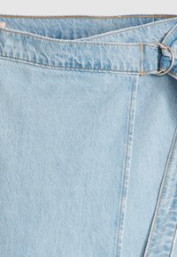 Jupe en denim bleu clair avec une large ceinture et une ceinture attachée maintenue par une boucle en D argentée sur fond blanc.