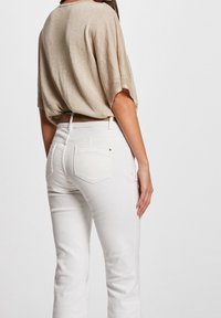 Beige kortærmet top med en tekstureret overflade, sat sammen med hvide tætsiddende jeans med baglommer og en subtil guld detalje på taljen.