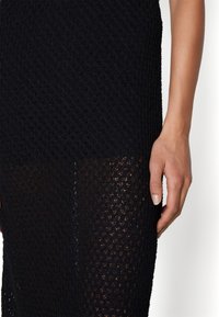 Robe en maille noire avec un motif texturé en losanges, présentant une partie supérieure ajustée et une section inférieure transparente. Comprend un gros plan sur une main.