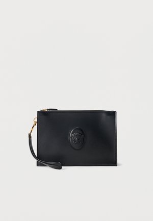 Pochette bracelet en cuir noir avec une fermeture éclair dorée et un emblème Méduse embossé au centre sur fond blanc.
