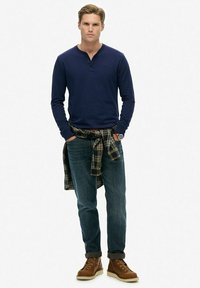Marineblauwe langemouwen henley shirt, blauwe jeans en bruine veterschoenen. Flanellen shirt om de taille geknoopt met een ruitpatroon.