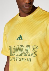 Κίτρινη αθλητική μπλούζα με πλέγμα και λαιμόκοψη. Διαθέτει το πράσινο λογότυπο "ADIDAS" και το κείμενο "SPORTSWEAR". Έχει υφή σε όλο το ύφασμα.
