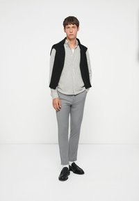 Camisa de botones a rayas grises debajo de un chaleco negro de punto, combinada con pantalones grises a medida y mocasines negros, sobre un fondo blanco liso.
