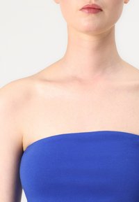 Zils strapless tops ar gludu tekstūru, cieši pieguļošu dizainu un platu joslu ap krūšu zonu. Fons ir neitrāls un gaišs.