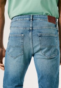 Jeans de mezclilla de un azul claro con un diseño clásico de cinco bolsillos, con una etiqueta de cuero marrón en la cintura y sutiles deslavados en la tela.