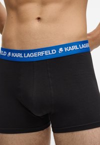 Vyrų liemuo vilkintis juodus apatiniai šortus su mėlyna juostele, ant kurios yra balta „KARL LAGERFELD“ užrašas ir logotipas.