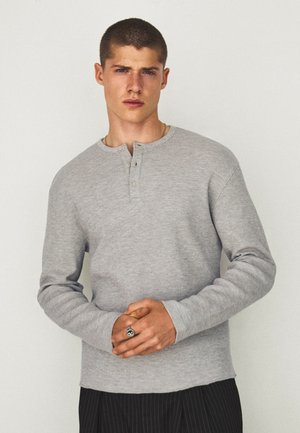 Giovane uomo con capelli corti che indossa una camicia a maniche lunghe grigio chiaro e pantaloni scuri a righe, in piedi con le mani giunte davanti.