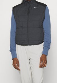 Sort puffer-vest med lynlås foran og høj krave, kombineret med en blå langærmet skjorte. Har sidelommer og letvægtsmateriale.