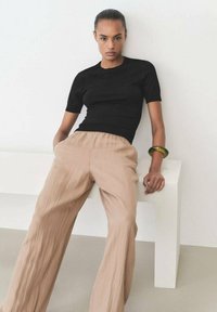 Black ribbed short-sleeve top paired with loose, textured beige trousers. The model wears a green bangle and stands against a neutral backdrop.