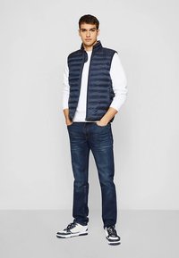 Marineblaue gesteppte Weste über einem weißen Pullover, kombiniert mit dunkelblauen Jeans und weißen Sneakers. Einfaches Design mit Reißverschluss und Seitentaschen.