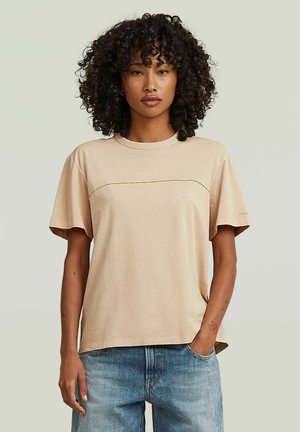Femme aux cheveux bouclés portant une chemise beige ample à manches courtes et un jean bleu, debout avec une main dans la poche devant un fond uni.