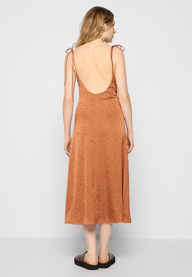 Vestido midi de satén naranja oxidado con tirantes ajustables, espalda descubierta y pequeños patrones de puntos en toda la prenda. Textura suave. Se llevan zapatos planos negros.