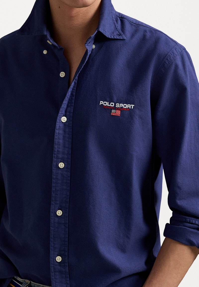 Camisa de botones azul marino de algodón, con un logo bordado "POLO SPORT" y un collar tipo doblado. Tejido texturizado con un acabado suave.