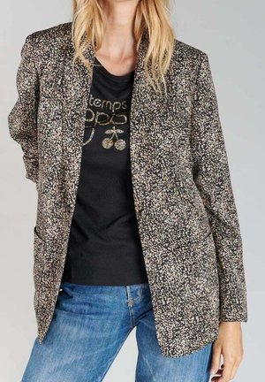 Mujer con una chaqueta marrón y beige con motas sobre una camiseta negra con diseño gráfico y vaqueros azules, de pie frente a un fondo liso.
