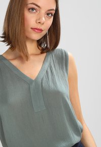 Haut sans manches dans une teinte bleu-vert atténuée, avec un décolleté en V, un tissu texturé et une coupe décontractée, agrémenté de détails subtils.