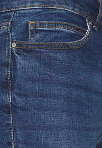 Jeans de mezclilla azul oscuro con un bolsillo trasero que presenta costuras naranjas y un detalle de remache. La tela tiene un acabado texturizado.