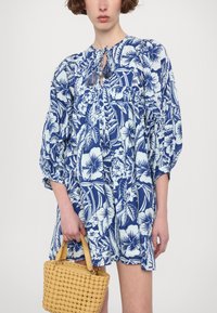 Blauwe bloemenjurk met pofmouwen en striknek, met een witte botanische print. Gecombineerd met een geweven gele handtas.