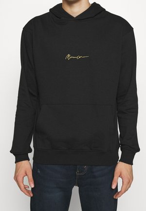 Mand iført en sort hættetrøje med gul kursiv tekst på brystet og mørkeblå jeans, stående mod en ensfarvet hvid baggrund.