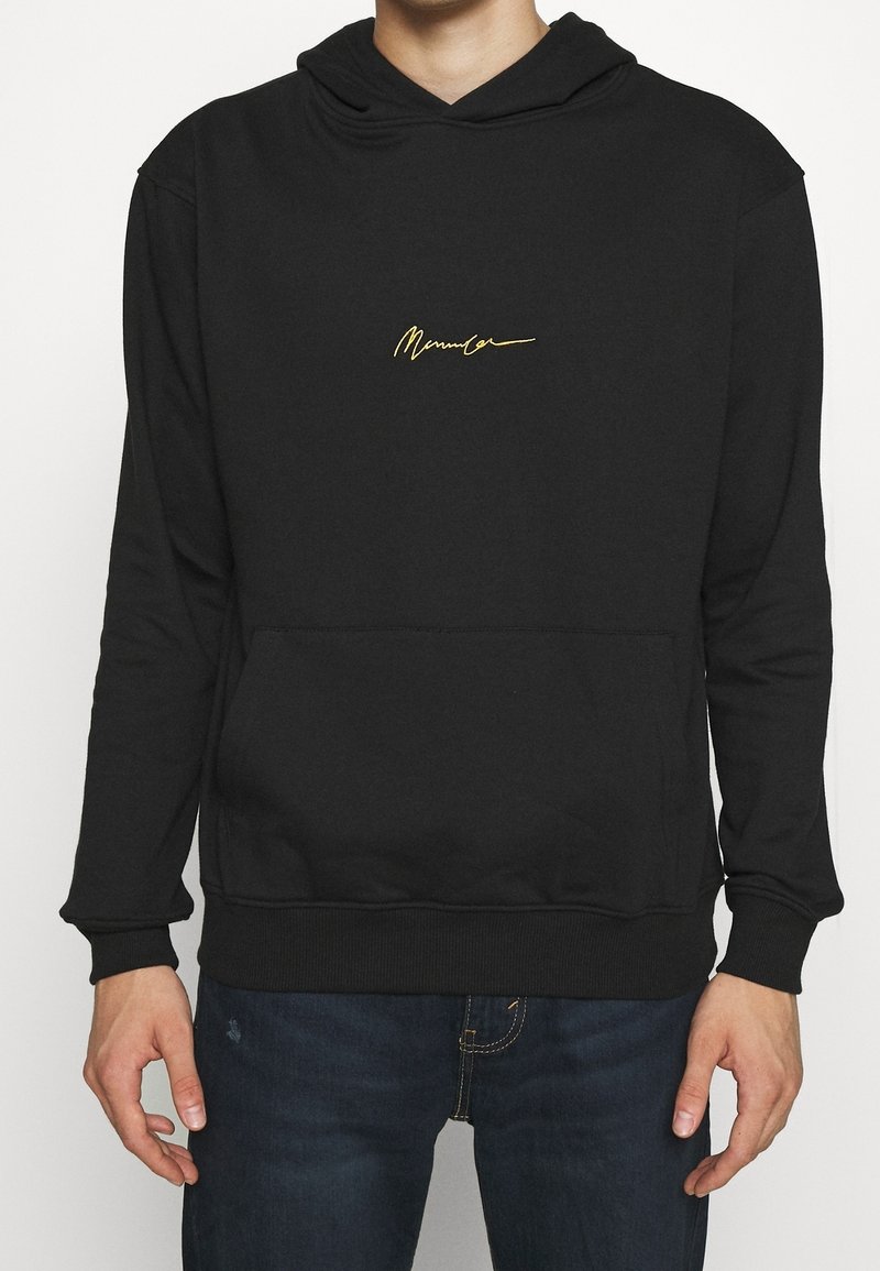 Homme portant un sweat à capuche noir avec un texte jaune en écriture cursive sur la poitrine et un jean bleu foncé, debout devant un fond blanc uni.