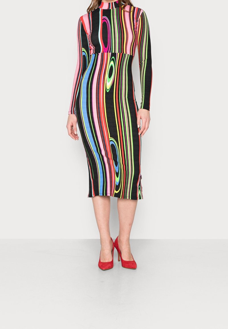 Femme portant une robe midi rayée colorée avec manches longues et des talons hauts rouges, debout contre un fond clair uni.
