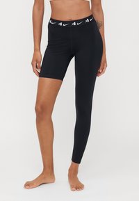 Juodos sportinės leggins su tampria medžiaga, turinčiomis kontrastingą juostą su Nike logotipu. Viena koja visiškai ilga, kita iki kelių.