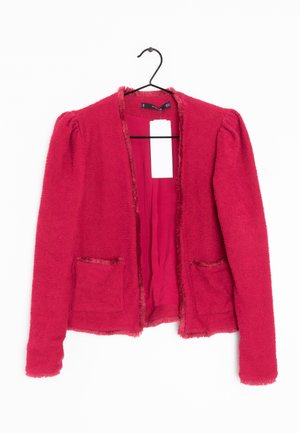 Veste femme rouge texturée à ouverture frontale, manches longues, deux poches avant et une étiquette blanche vierge suspendue à un cintre noir.