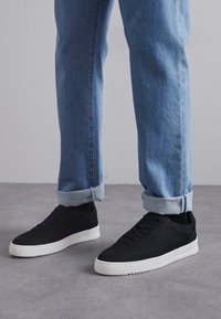 Sorte semskede sneakers med hvite gummisåler, med runde lisser og et slankt, minimalistisk design. Brukt med rullet lyseblå denimjeans.