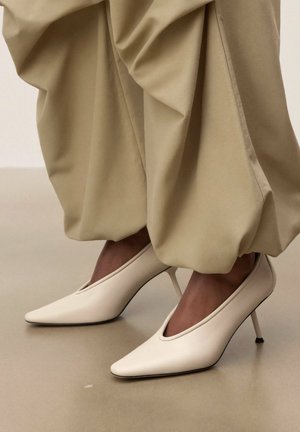 Nohy v bielych vysokých podpätkoch so špicatou špičkou, v kombinácii s voľnými béžovými nohavicami stiahnutými pri členkoch na neutrálnom pozadí.