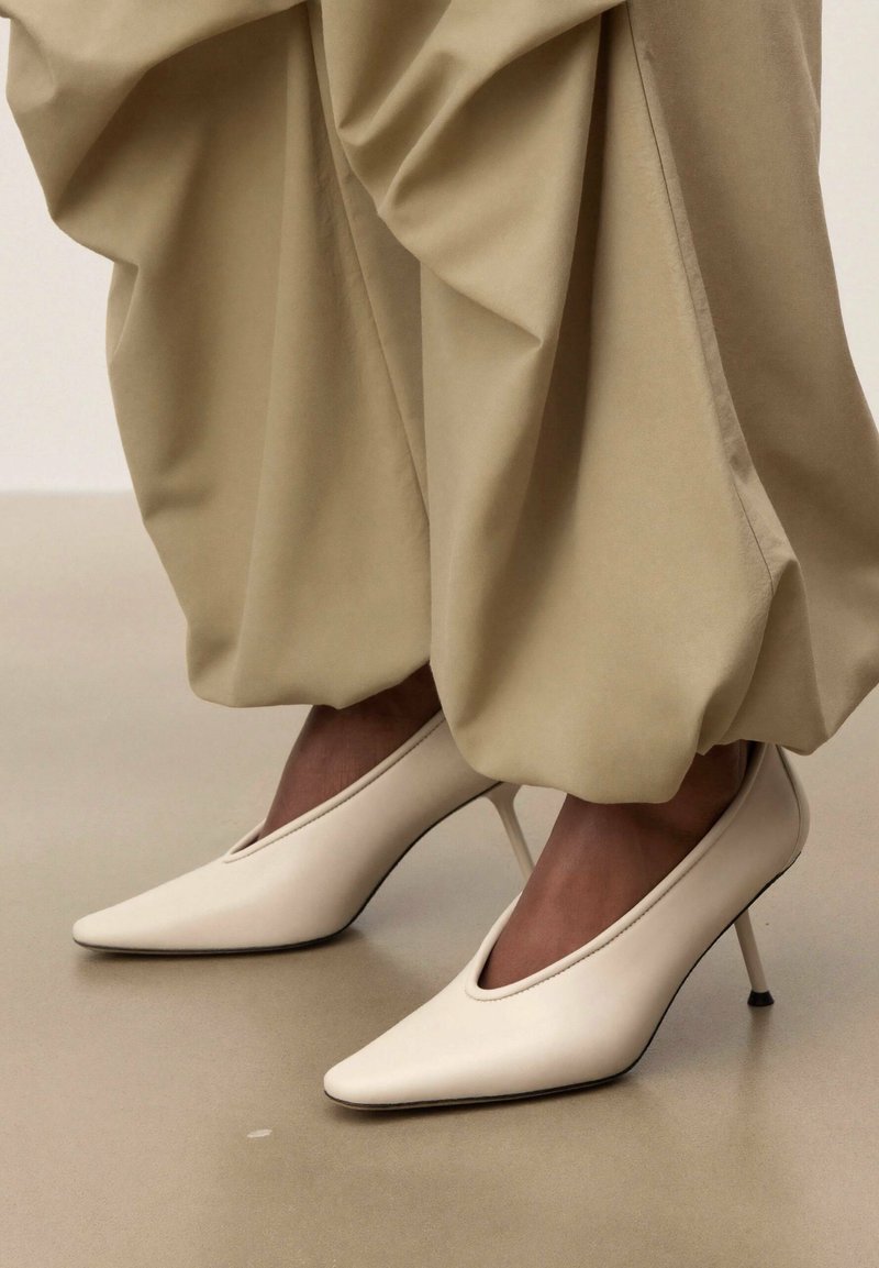 Pieds chaussés de talons hauts blancs à bouts pointus, associés à un pantalon beige ample resserré aux chevilles, sur un fond neutre.