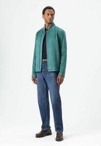 Turquoise suède jas met een opstaande kraag, gecombineerd met blauwe spijkerbroek en donkerbruine schoenen. Glad oppervlak en strakke lijnen.