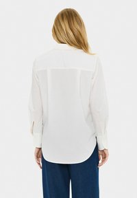 Donna con lunghi capelli biondi che indossa una camicia bianca a maniche lunghe e pantaloni blu scuro, in piedi di spalle rispetto alla macchina fotografica.