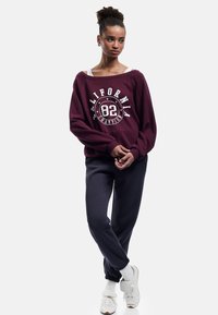 Sudadera oversized color burdeos con estampado gráfico y logo en blanco, combinada con pantalones de chándal oscuros azul marino y zapatillas blancas. Fondo liso.