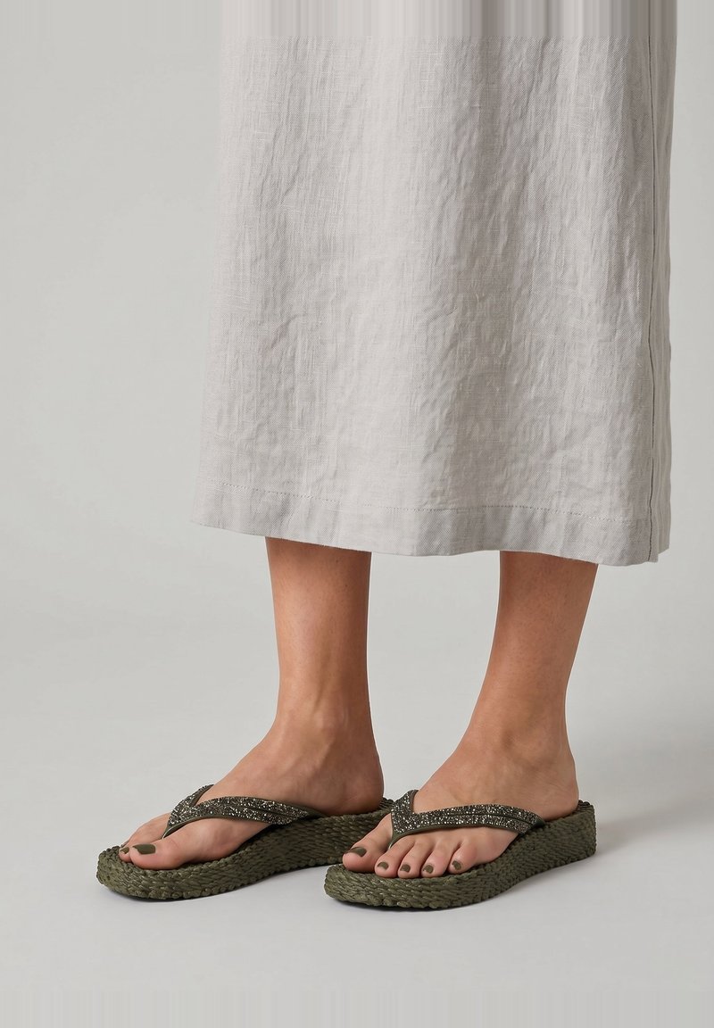 Pies con chanclas de plataforma verdes oliva con tiras texturizadas, de pie sobre una superficie clara, vestidos con un vestido de color claro por debajo de las rodillas.