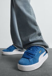 Sinised Puma sametist tossud paksude paeltega, kantud vabalt istuvate helesiniste teksadega hallil põrandal.