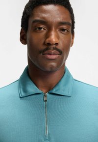 Turkusowa polo z zamkiem błyskawicznym, wykonana z teksturowanej tkaniny, z kołnierzykiem, wyposażona w metalowy zamek i proste, czyste linie.