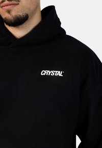 Černá mikina s kapucí vyrobená z měkké tkaniny, s přední kapsou a výrazným bílým logem "CRYSTAL" na hrudi. Kapuce s nastavitelným stahováním.