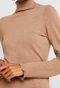 Pull col roulé beige en tissu doux et texturé avec manches longues et poignets à volants. Présente une silhouette ajustée et un motif côtelé subtil.