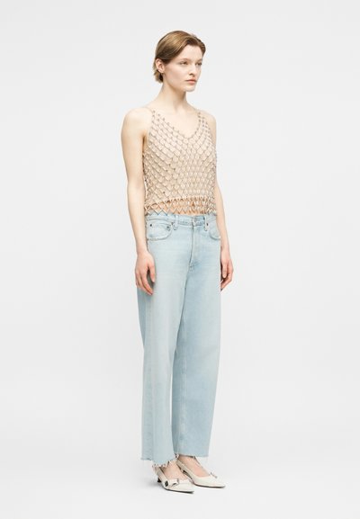 Γυναίκα που φοράει μπεζ διαφανές αμάνικο τοπ, ανοιχτό μπλε τζιν παντελόνι φαρδύ με ξεφτισμένα τελειώματα και λευκά ψηλοτάκουνα παπούτσια μυτερά με λεπτομέρεια αγκράφας.