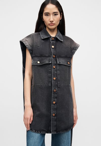 Νεαρή γυναίκα που φοράει αμάνικο μαύρο τζιν γιλέκο με κουμπιά, μεγάλες τσέπες στο στήθος και ξεθωριασμένες λεπτομέρειες, σε συνδυασμό με μπλε τζιν.